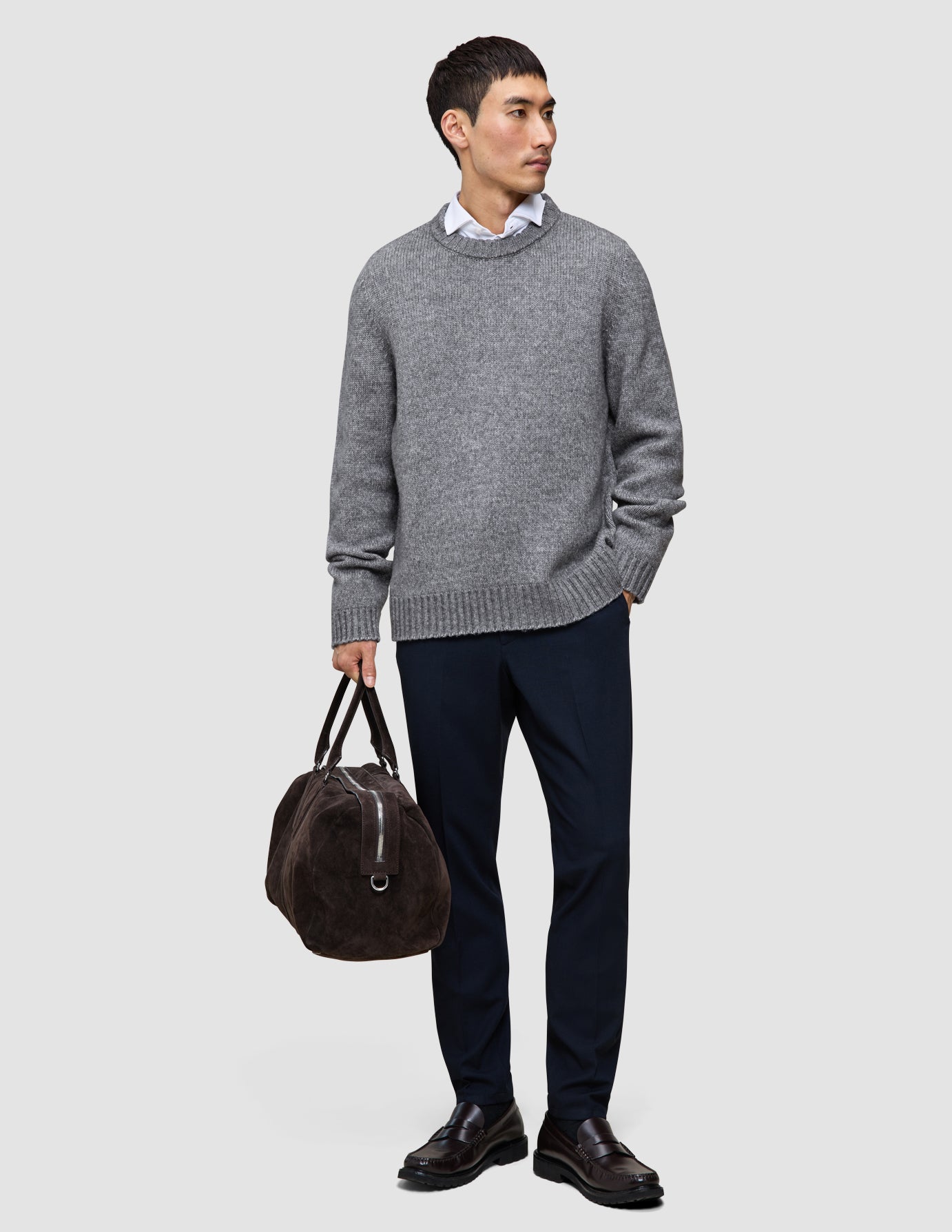 A man wears a Chunky Soft Wool Knit Crewneck Glacier Grey sweater over a white collared shirt, dark trousers, and dark loafers, while holding a brown duffel bag.